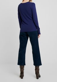 Top de manga larga azul marino con escote redondeado y puños abotonados, combinado con pantalones de pana oscuros de pierna ancha y botas marrones con patrón.