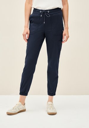 Vrouw draagt marineblauwe joggingbroek met trekkoord en elastische boorden en witte sneakers, staand tegen een effen achtergrond.