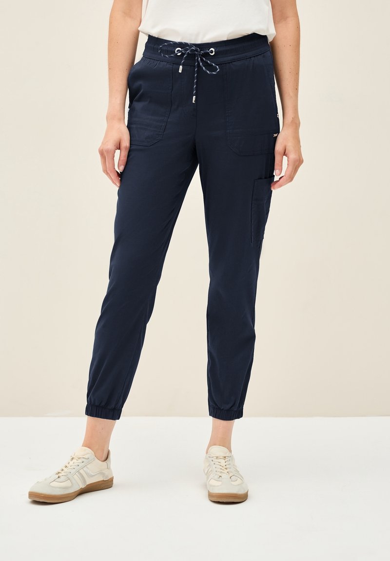 Femme portant un pantalon de jogging bleu marine avec cordon de serrage et poignets élastiques ainsi que des baskets blanches, debout devant un fond uni.
