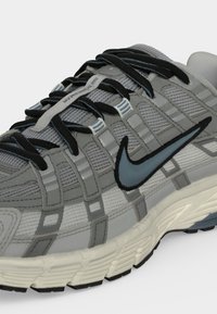 Γκρι αθλητικό παπούτσι Nike με μαύρα κορδόνια, μπλε λογότυπο Swoosh, ανθεκτικό αεριζόμενο πλέγμα και λευκή αντιολισθητική ενδιάμεση σόλα με μαύρη εξωτερική σόλα.