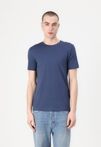 Μπλε ναυτικού κοντομάνικο μπλουζάκι φτιαγμένο από μαλακό ύφασμα, με στρογγυλή λαιμόκοψη και εφαρμοστή σχεδίαση. Φοράει ελαφριά μπλε τζιν.