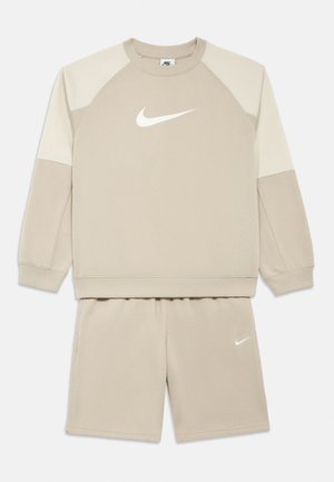 Beżowy dres marki Nike, składający się z bluzy i dopasowanych szortów z białymi logo Nike, długimi rękawami oraz kontrastującymi jaśniejszymi panelami na ramionach bluzy.
