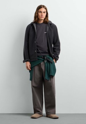 Young person with shoulder-length hair wears black fleece jacket, dark t-shirt, wide gray pants, green sweater tied at waist, and beige shoes.