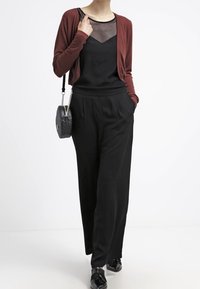 Schwarze weite Hosen, schwarzes Tanktop mit Mesh-Detail und eine burgunderfarbene Strickjacke. Die Accessoires umfassen eine runde schwarze Tasche und glänzende schwarze Schuhe.