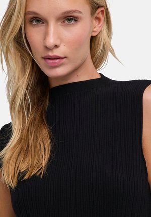 Blonde vrouw met groene ogen, gekleed in een zwart geribd mouwloos topje, kijkt iets van de camera weg tegen een effen achtergrond.