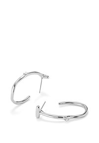 Boucles d'oreilles en argent avec un design élégant et cintré, agrémentées d'une petite pierre en haut. La texture est lisse et polie.