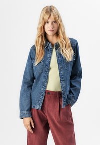 Woman with long blonde hair wearing an unbuttoned denim jacket, light yellow top, and high-waisted rust-colored corduroy pants.