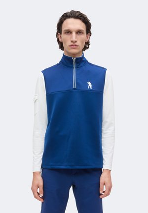 Homme debout face à l'avant, portant un gilet bleu sans manches à fermeture éclair sur une chemise blanche à manches longues et un pantalon bleu, sur fond uni.
