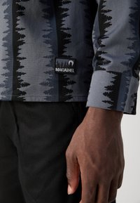 Camisa de manga longa com um padrão de árvore em cinza e preto, com uma etiqueta de tecido a dizer "MAKIA&HEL." O material parece liso e durável.