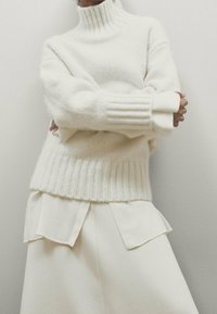 Pull en tricot crème avec un col haut à côtes et de larges poignets côtelés. Superposé sur une blouse blanche avec des rabats pointus. Texture douce.