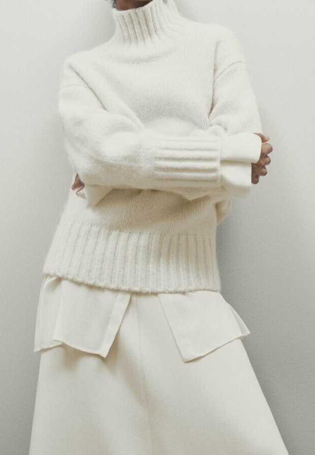 Pull en tricot crème avec un col haut à côtes et de larges poignets côtelés. Superposé sur une blouse blanche avec des rabats pointus. Texture douce.