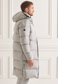 Abrigo largo acolchado gris con capucha, que presenta un diseño acolchado, cierre de cremallera y un acento negro en la manga. Textura suave en todo.