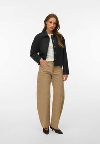 Femme portant une veste noire boutonnée, un haut blanc, un pantalon large couleur sable et des talons noirs, debout avec une main dans la poche.