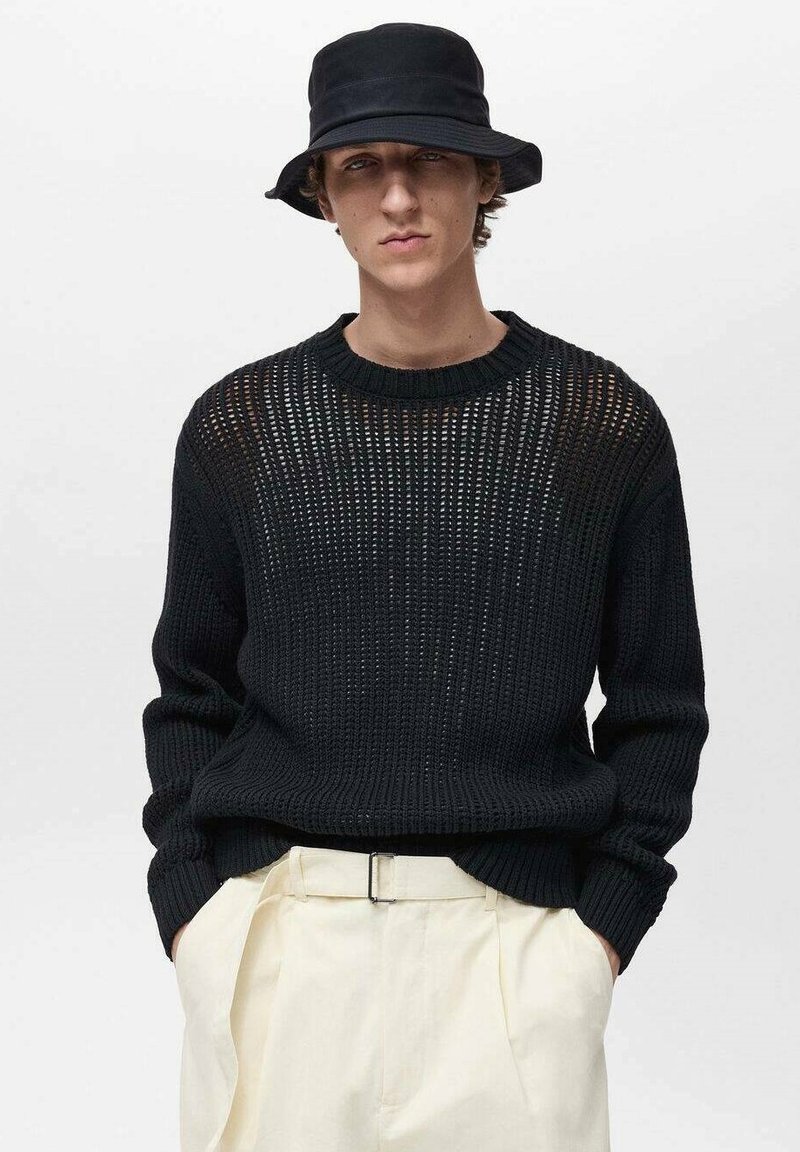 Schwarzer Strickpullover mit offener Maschenstruktur, Rundhalsausschnitt und gerippten Bündchen, kombiniert mit einem schwarzen Bucket Hat und cremefarbenen Hosen.