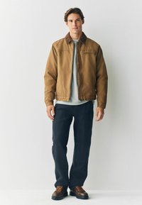 Brown leather jacket with a corduroy collar, gray sweater, dark pants, and brown boots with orange laces, standing against a light background.