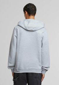 Uomo che indossa una felpa grigia chiara con cappuccio vista da dietro, abbinata a pantaloni cargo neri su uno sfondo grigio semplice.