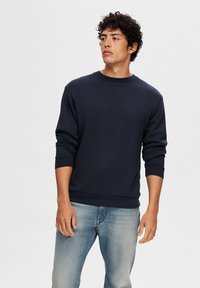 Sudadera marina de algodón, con cuello redondo, manga larga, y puños y dobladillo acanalados, combinada con jeans de color azul claro.