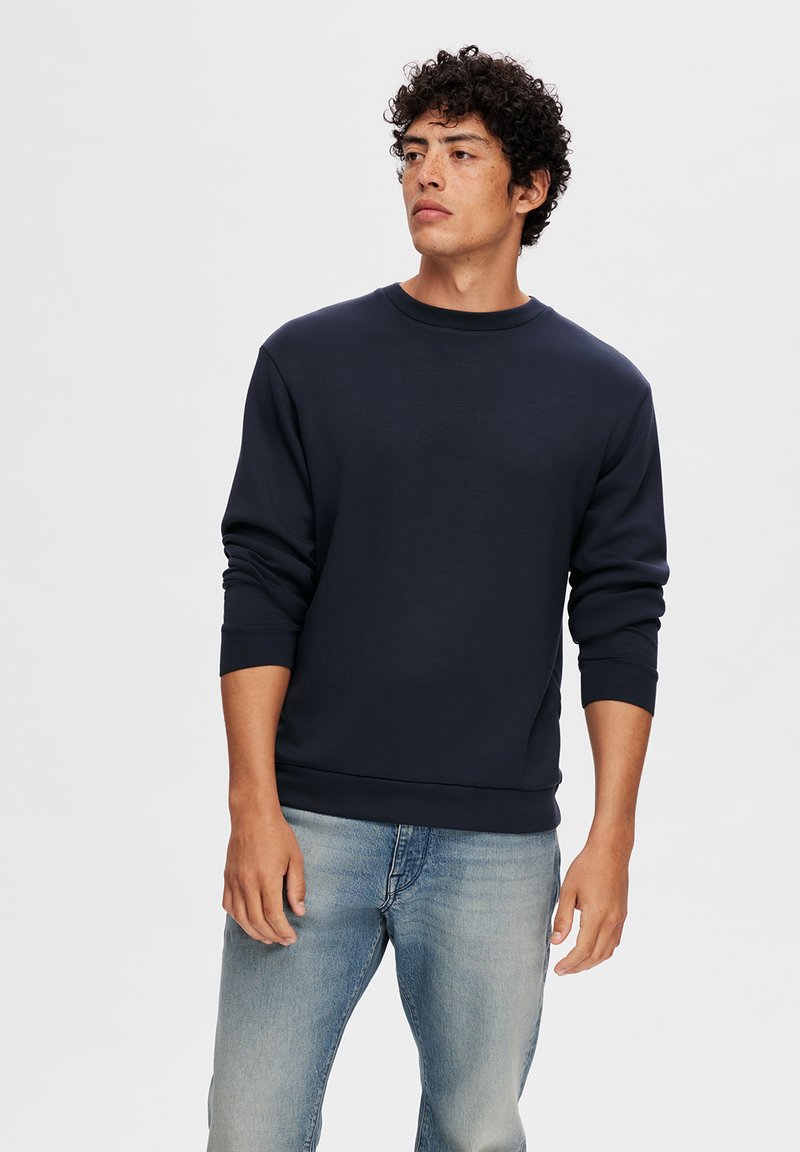 Sudadera marina de algodón, con cuello redondo, manga larga, y puños y dobladillo acanalados, combinada con jeans de color azul claro.