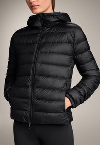 Črn puhasti jopič z horizontalnim šivanim dizajnom, z zadrgo spredaj in pritrjeno kapuco. Mehak, gladek material z elastičnimi manšetami.