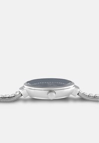 Silberfarbene Armbanduhr mit rundem Gehäuse, dunklem Zifferblatt und Kristallakzenten. Das flexible Metallband zeichnet sich durch ein strukturiertes, gewebtes Design aus.