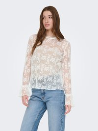 Blouse à manches longues en dentelle ivoire avec broderies florales, encolure festonnée et poignets à volants. Associée à un jean en denim bleu clair.