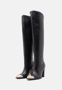 Botas de cuero negras sobre la rodilla con un acabado suave, que presentan una puntera beige y un tacón cuadrado. Costuras detalladas a lo largo del borde superior.