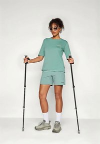 Gaiši zaļš aktīvo apģērbu komplekts, kas sastāv no īsām piedurknēm un šortiem. Aksesuāri ietver pārgājienu nūjas un pelēkas sporta kurpes ar baltām zeķēm.