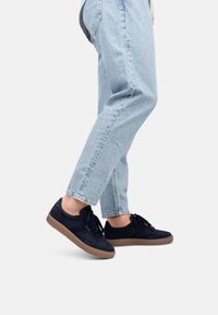 Zapatillas bajas de ante azul marino con cordones oscuros y suela de goma marrón, combinadas con jeans de pierna recta de un azul claro.