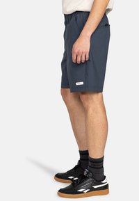 Mörkblå shorts med elastisk midja, sidofickor och en etikett vid fållen. Bärs med svarta sneakers och grå strumpor.