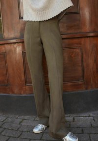 Olivgrüne, ausgestellte Hose aus glattem Stoff, mit vorderen Nähten und einer taillierten Passform, kombiniert mit silbernen Sneakers.