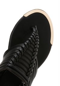 Black leather sandal featuring a braided design, flat sole, and a gold accent at the toe. Smooth texture with minimal hardware details.