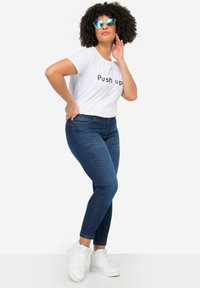 Camiseta blanca con el texto "Push up", jeans ajustados azules y zapatillas blancas. El modelo posa con confianza, mostrando un atuendo casual.