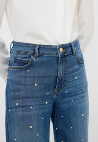 Jeans z wysokim stanem w niebieskim kolorze, z przednimi kieszeniami, złotym zapięciem na guzik oraz rozproszonymi ozdobami w kształcie pereł z przodu.