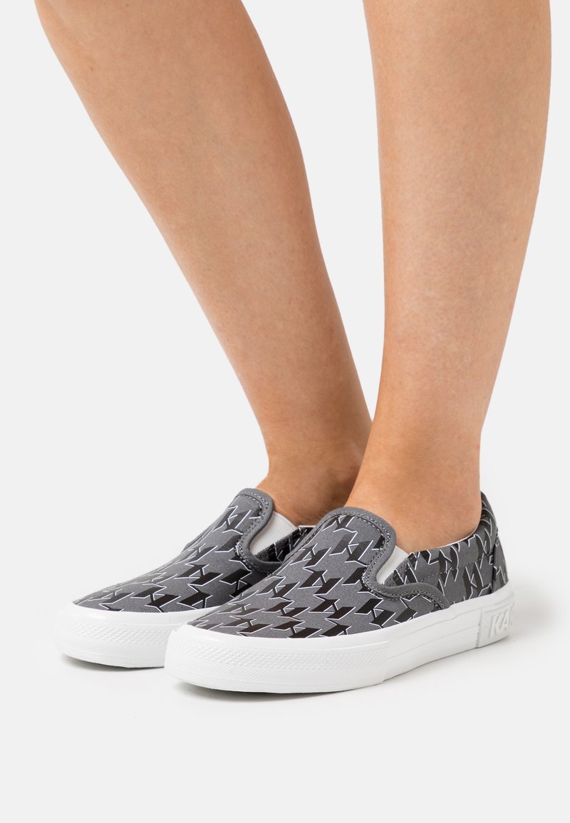 Paire de pieds portant des baskets grises sans lacets avec semelles blanches et motif étoile géométrique noire sur fond clair.