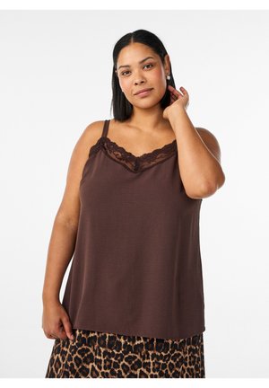 Žena v hnedom tielku s čipkovou lemovkou a sukni s leopardím vzorom, dotýka sa krku, pózuje na bielom pozadí.