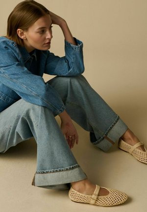 Femme assise par terre portant une veste en jean bleue et un jean à jambes larges avec des ballerines Mary Jane tissées beiges sur un fond beige uni.