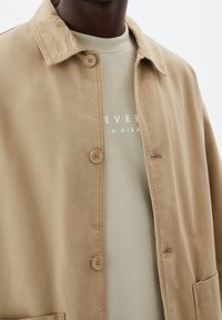 Beige bomuldsjakke med krave, to knapper og sidelommer båret over en lysegrøn sweatshirt med hvid tekst.