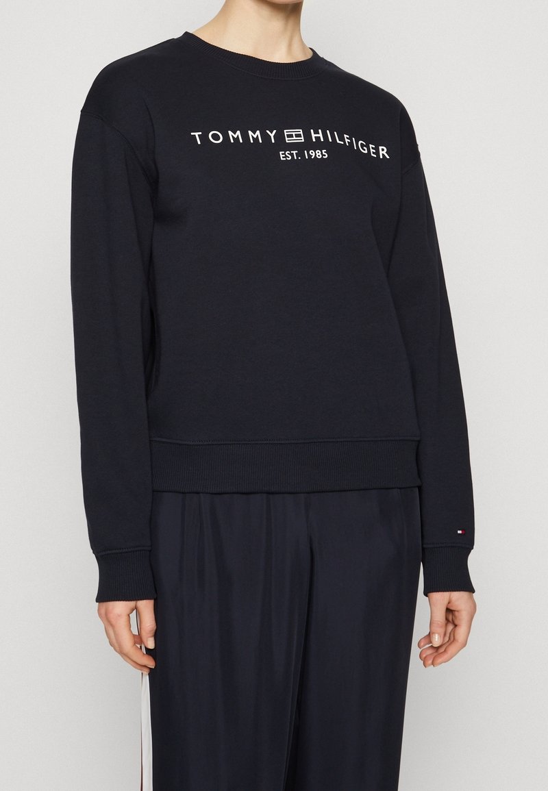 Personne portant un sweat-shirt noir Tommy Hilfiger avec un logo blanc et le texte "EST. 1985" sur la poitrine et un pantalon noir assorti.