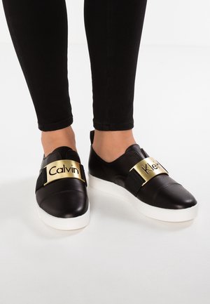 Zapatillas negras tipo slip-on con suelas blancas y tiras metálicas doradas con la inscripción "Calvin Klein" en pies que llevan leggings negros.