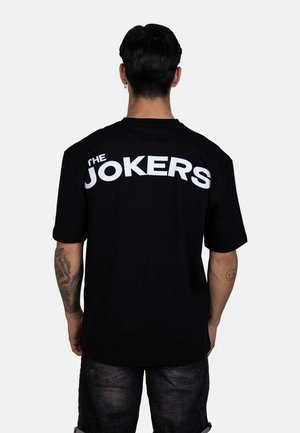 Hombre con cabello corto y oscuro de espaldas, vistiendo una camiseta negra con "THE JOKERS" en grandes letras blancas en la espalda.