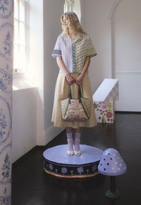 Kobieta w kwiecistej bluzce z patchworku i beżowej spódnicy trzyma pasującą kwiecistą torbę, stojąc na udekorowanej okrągłej platformie obok dużego fioletowego modelu grzyba.