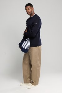Suéter azul marino con acentos de botones en el hombro, chinos beige y zapatillas de color blanco roto. Bolsa a rayas en azul marino y blanco sostenida en el brazo izquierdo.