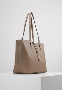 Beige nahka-tote-pussi, jossa on kaksi pitkää kahvaa, minimalistinen muotoilu ja ulkopuolella oleva vetoketjusulku, jossa on koristeellinen vetoketjun yksityiskohta.