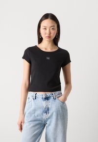 Black cropped t-shirt with short sleeves and a round neckline, featuring a small logo patch at the center. Paired with light-wash denim jeans.