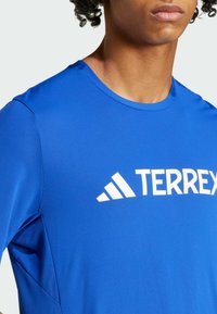 Camiseta deportiva de poliéster azul con cuello redondo y logo blanco "TERREX", con una textura suave y mangas cortas.