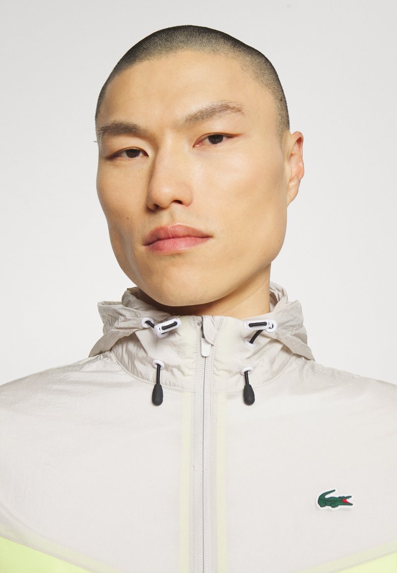 Lichte, gewatteerde jas met capuchon in lichtgrijs met gele accenten. Voorzien van een rits, trekkoorden en een klein geborduurd logo. Goederen textuur.