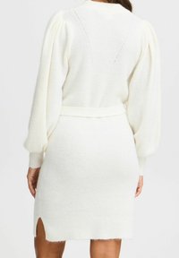 Robe pull en blanc doux, présentant des manches bouffantes, une taille ceinturée et des fentes sur le côté à l'ourlet. Tissu texturé et douillet.