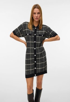 Robe tricotée à manches courtes en noir et blanc à carreaux avec des boutons, deux poches poitrine et un ourlet côtelé. Portée avec des bottes noires arrivant au genou.