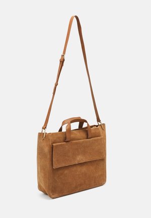 Bolso de hombro de ante marrón con bolsillo delantero con solapa, asas dobles y correa de cuero ajustable sobre un fondo liso.