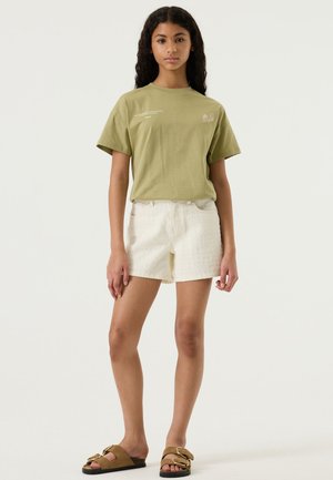 Jonge vrouw die een olijfgroen T-shirt, witte gestructureerde short en bruine sandalen draagt, staand tegen een eenvoudige lichte achtergrond.